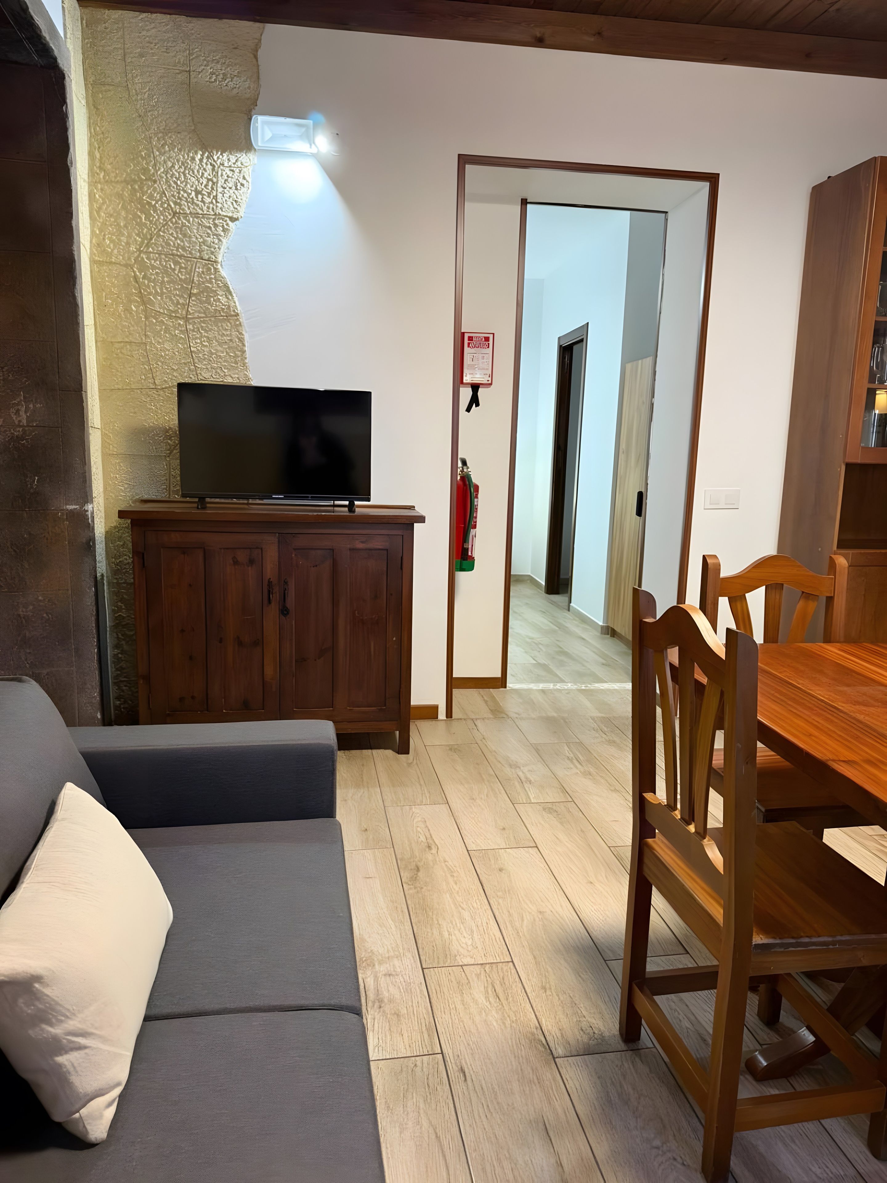 Een woonkamer met een flatscreen televisie op een houten kast, een grijze bank en een houten eettafel met stoelen.