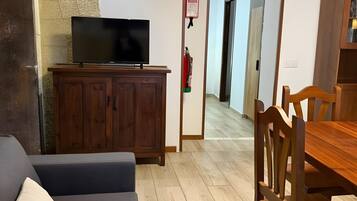 Een woonkamer met een flatscreen televisie op een houten kast, een grijze bank en een houten eettafel met stoelen.