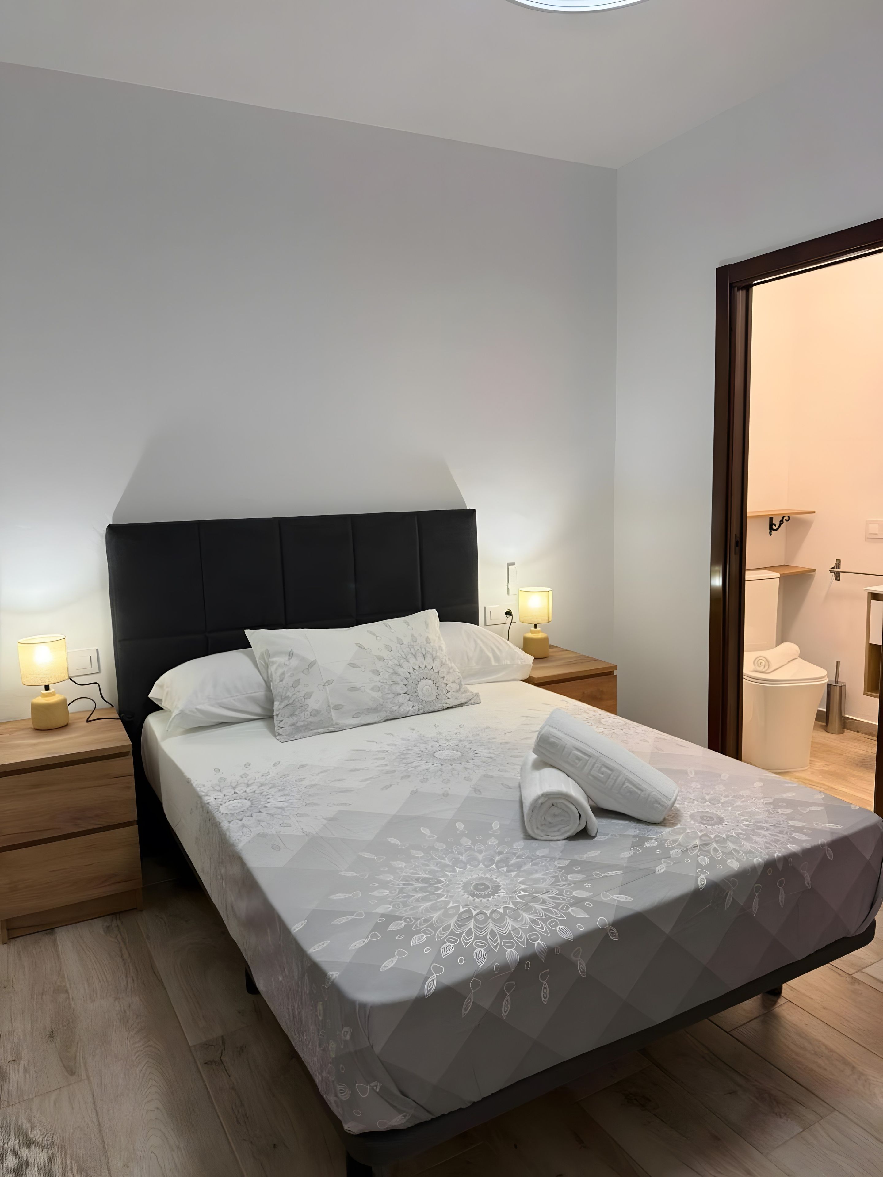 Una cama bien hecha con una colcha estampada, dos lámparas de noche, una mesita de noche de madera y vistas al baño a través de una puerta abierta.