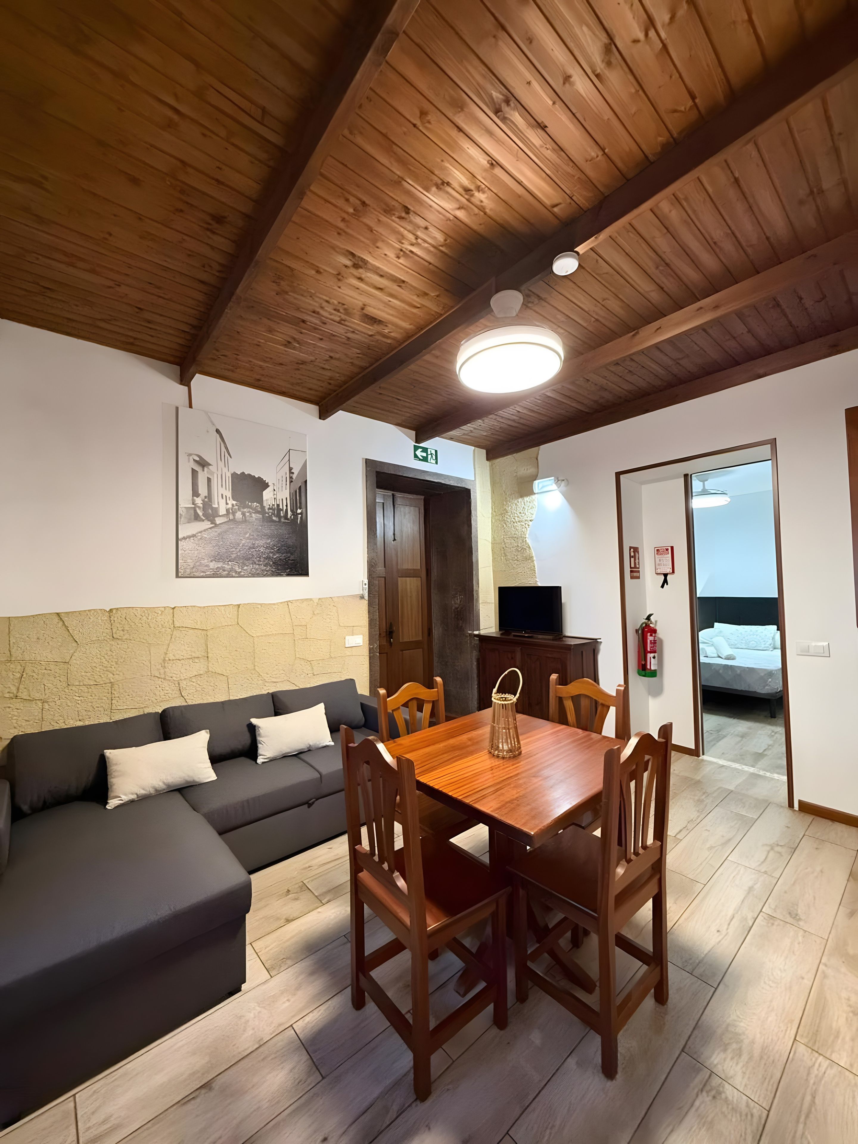A room with a wooden ceiling, a sofa, a dining table with chairs, a television, and a bed.