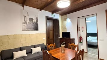 A room with a wooden ceiling, a sofa, a dining table with chairs, a television, and a bed.