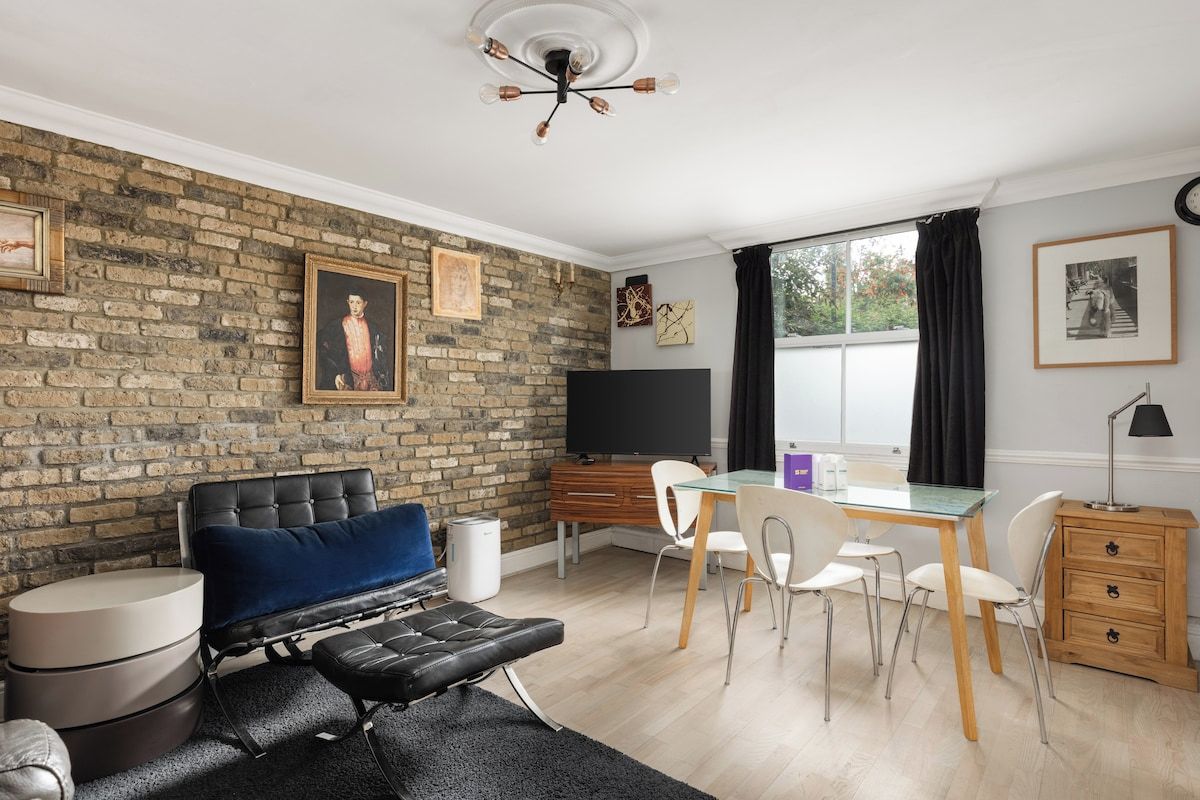 A modern living room with a brick wall, a blue sofa, a dining table with chairs, a TV, and a wooden cabinet.