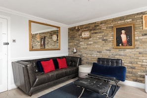 A room with a brick wall, a large mirror, a leather sofa with red cushions, a blue chair, and a small round table. - New! Stylish 2BR Flat near Brockwell Park (Greater London)