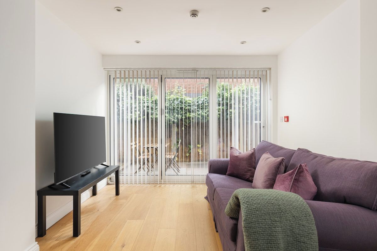 A modern living room with a flat-screen TV on a black stand, a purple sofa with cushions, and a sliding glass door leading to an outdoor area.