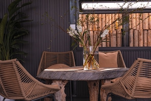 Een ronde houten tafel met twee rieten stoelen, een vaas met takken en witte bloemen, en een achtergrond van verticale houten latten.