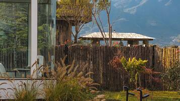 Una valla de madera rústica con una estructura de techo de paja, un edificio de cristal con una mesa y sillas, y un paisaje montañoso al fondo.