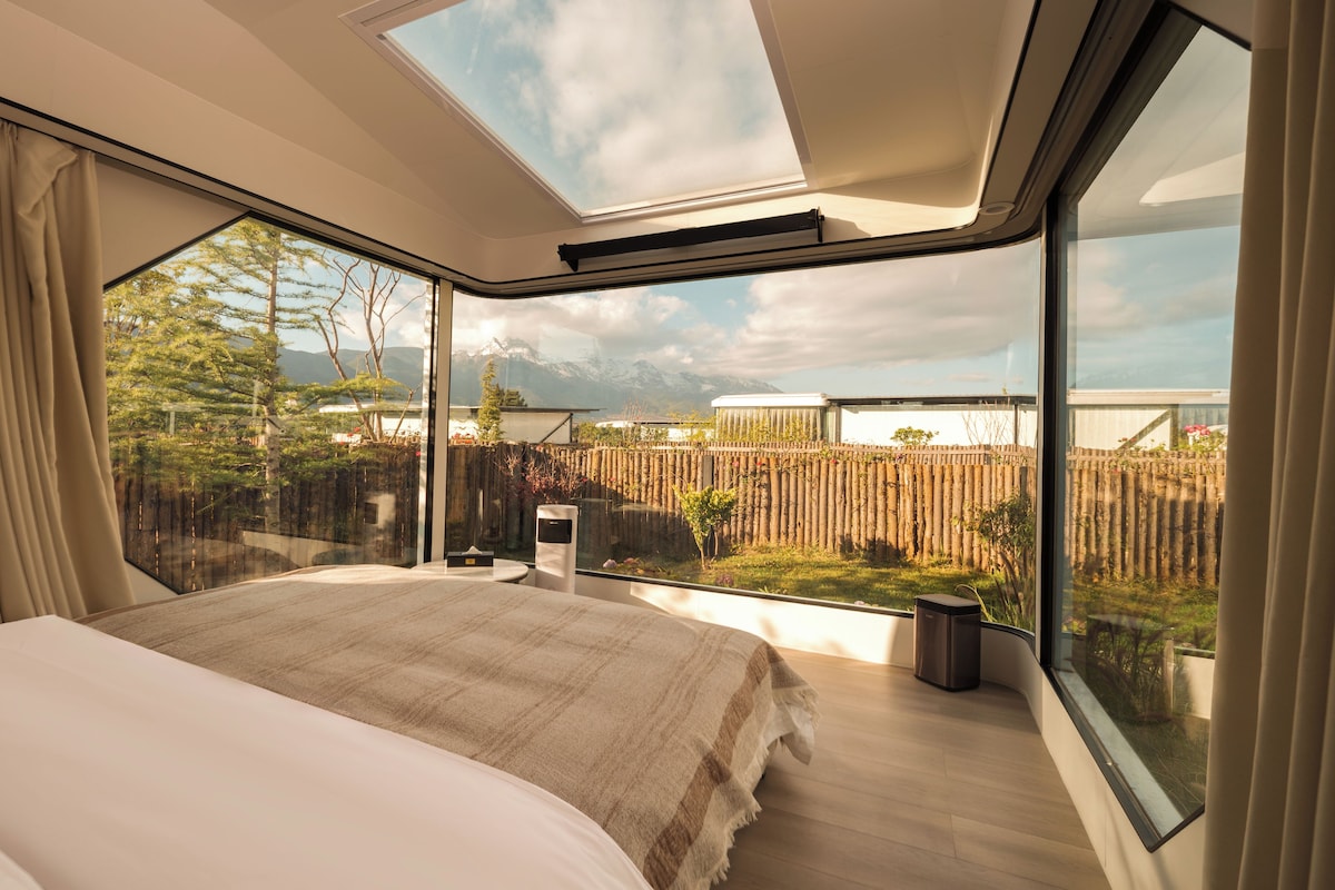 Een slaapkamer met een groot bed, dakraam en uitzicht op een tuin en bergen.