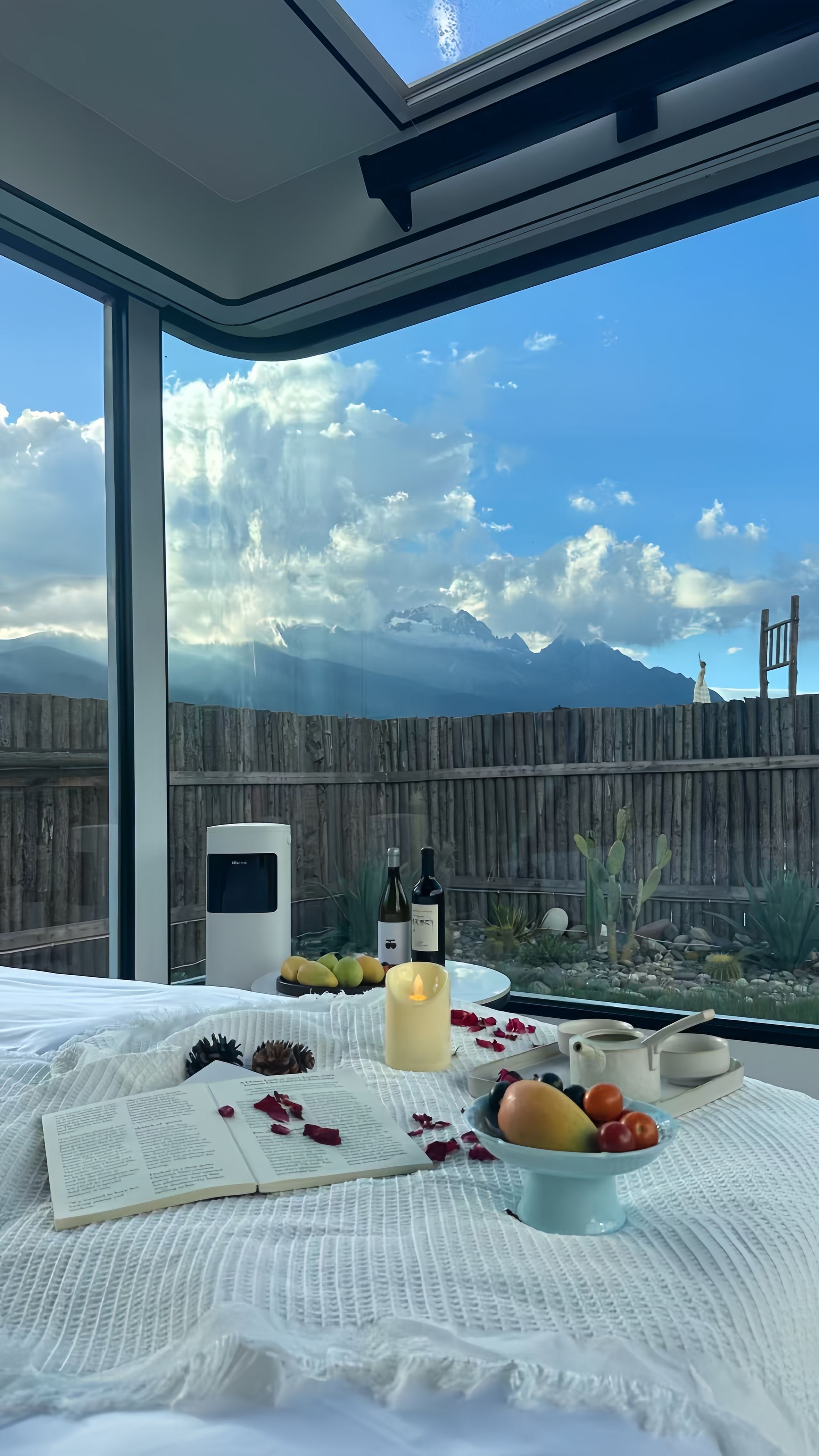 A bedroom with a large window overlooking a mountainous landscape, a bed with a white comforter, a bedside table with a candle, a bowl of fruit, and a wine bottle.