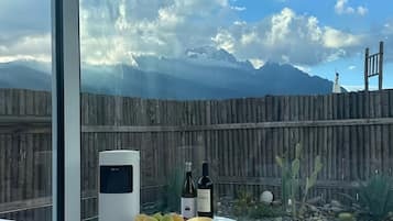 A bedroom with a large window overlooking a mountainous landscape, a bed with a white comforter, a bedside table with a candle, a bowl of fruit, and a wine bottle.