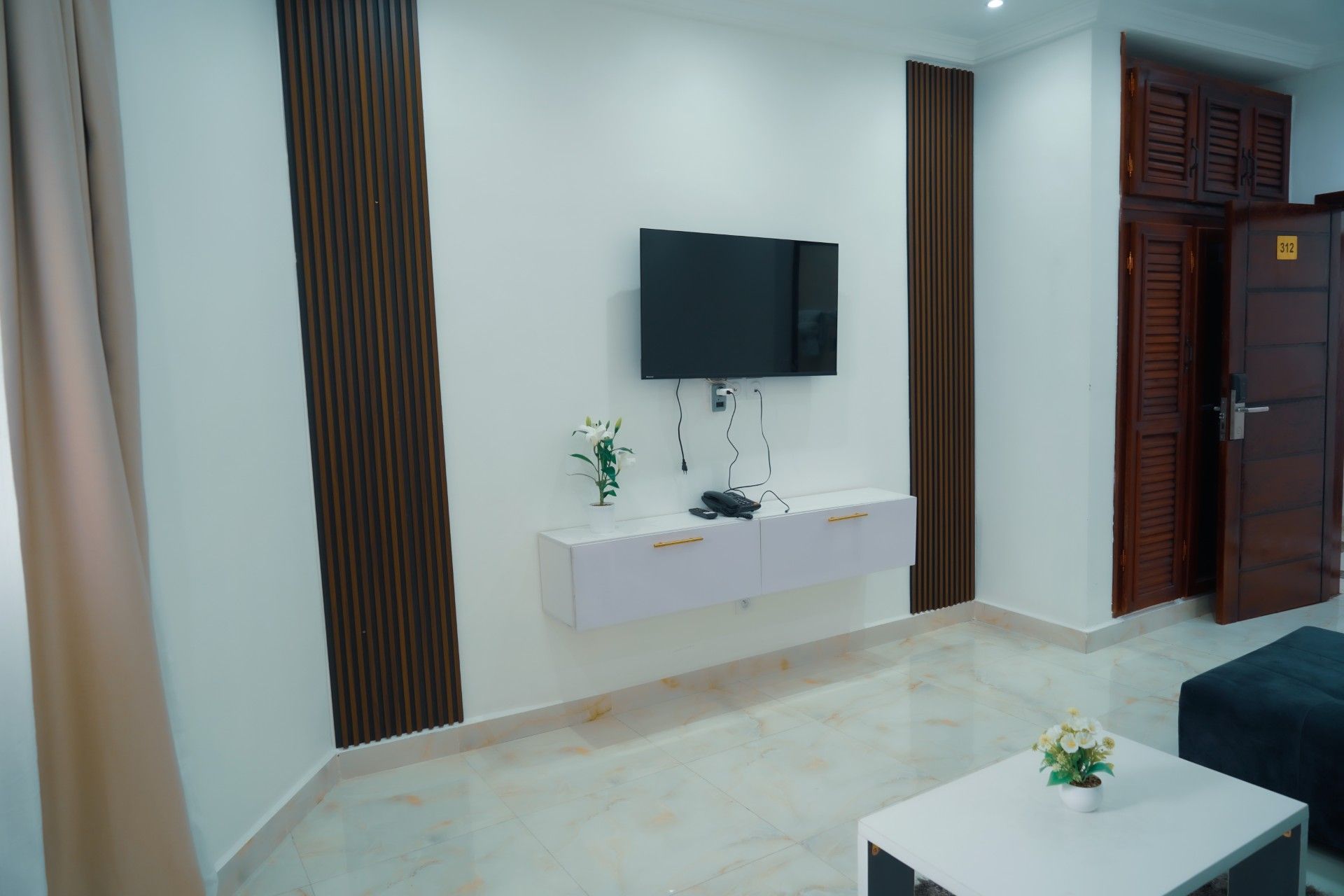 Una sala moderna con un televisor de pantalla plana montado en la pared, una mesita blanca con un teléfono y una pequeña planta en maceta.