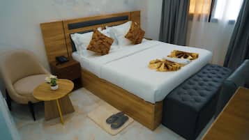 Habitación de hotel con cama, mesita de noche con teléfono, una silla, una pequeña mesa con una planta y un par de zapatillas en el suelo.