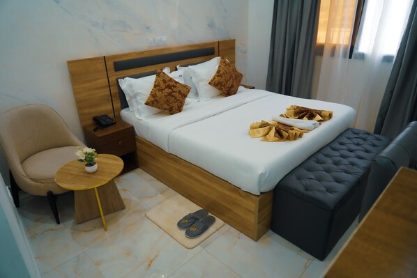 Habitación de hotel con cama, mesita de noche con teléfono, una silla, una pequeña mesa con una planta y un par de zapatillas en el suelo.