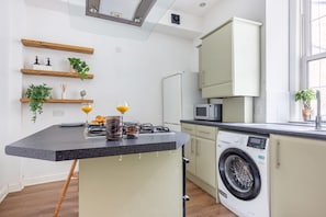 A compact kitchen with a built-in washing machine, a microwave, and wooden shelves with plants. - Spacious Victorian 2BR by Leith Walk (Edinburgh)