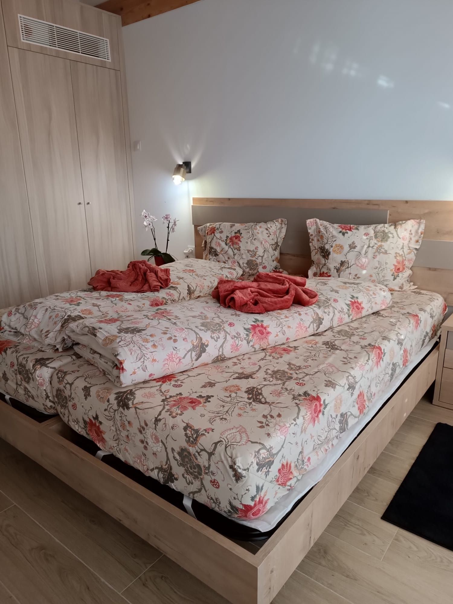 A bedroom with a wooden bed frame, floral bedding, a nightstand, and a wall-mounted light fixture.