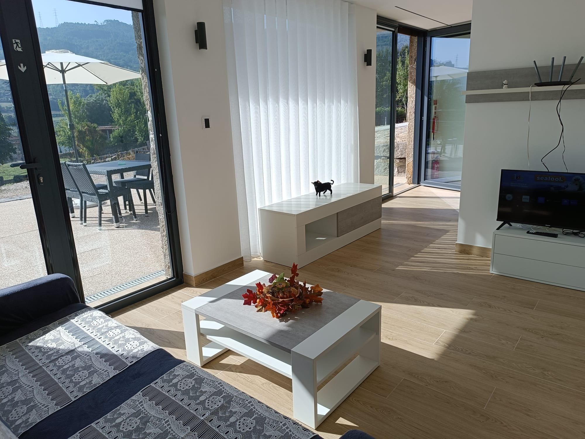 A modern living room with a sofa, a coffee table with a decorative centerpiece, a television, and a sliding glass door leading to an outdoor area with a table and chairs.