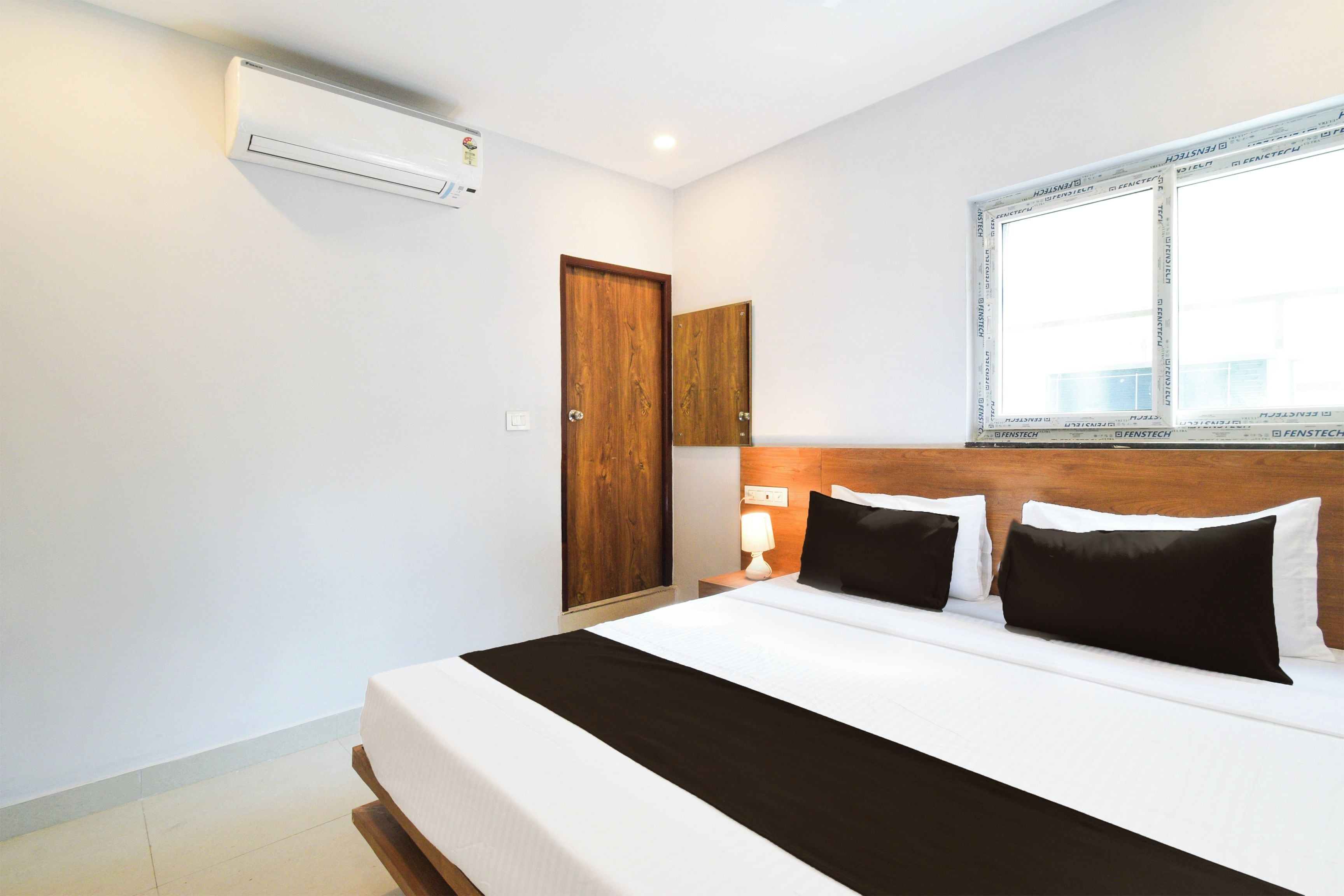 A hotel room with a bed, a wooden wardrobe, a nightstand with a lamp, and an air conditioning unit mounted on the wall.