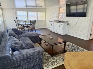 A living room with a sofa, a coffee table, a dining table with chairs, a kitchen area with cabinets, and a TV mounted on the wall. - You can bring everybody, have your own rooms, and still actually enjoy being together. (Montgomery)
