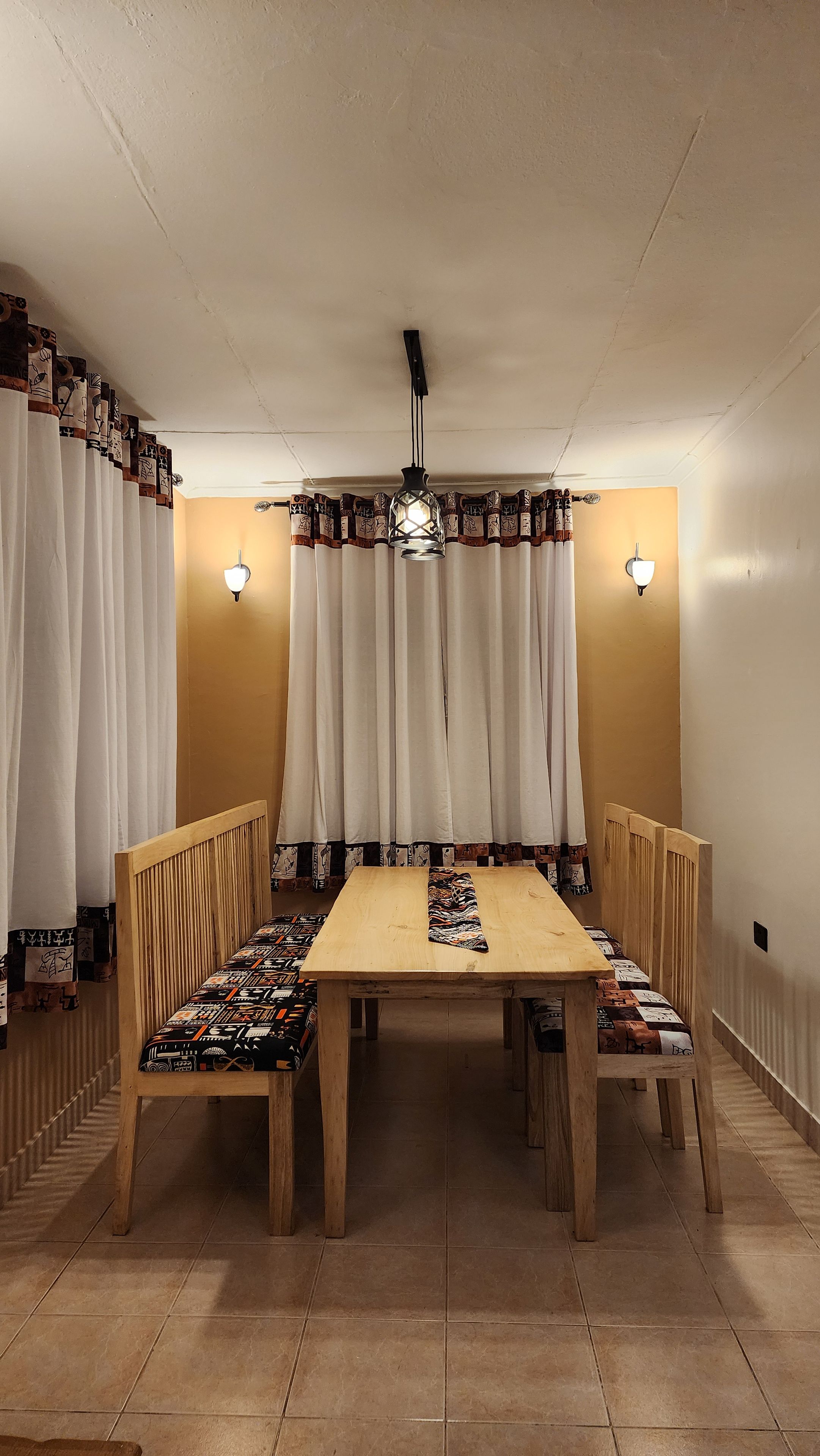 A dining area with a wooden table and chairs, white curtains, and wall-mounted lights.