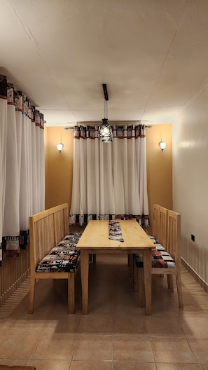 A dining area with a wooden table and chairs, white curtains, and wall-mounted lights. - Baiskeli Experience Haus | Cosy Arusha Stay for 6, Near Clock Tower (Arusha)