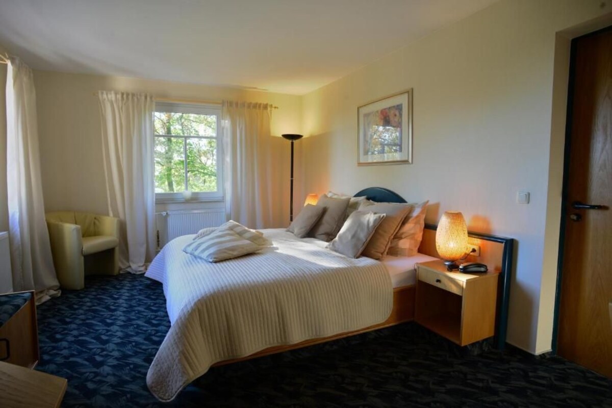 Een hotelkamer met een bed, nachtkastje, lamp, stoel en een raam met gordijnen.