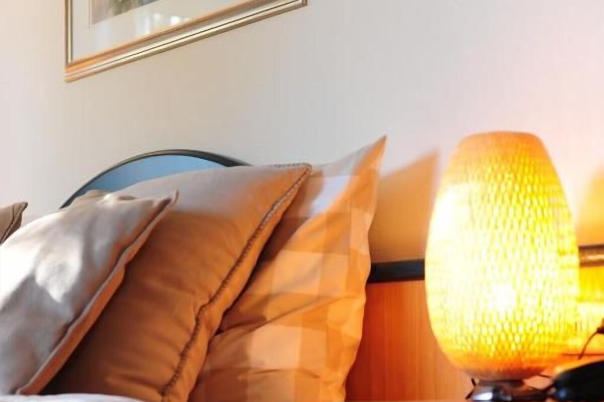 Een bed met een kussen, een nachtkastje met een lamp en een ingelijst beeld aan de muur.