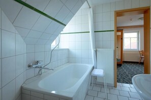Een badkamer met een bad, een douche en een toilet. Er is een raam dat voor daglicht zorgt en een deur die naar een andere kamer leidt.