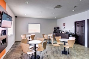 A modern interior with multiple round tables and chairs, a kitchen area, and a large window with blinds. - Motel 6 Innisfail AB (Innisfail)