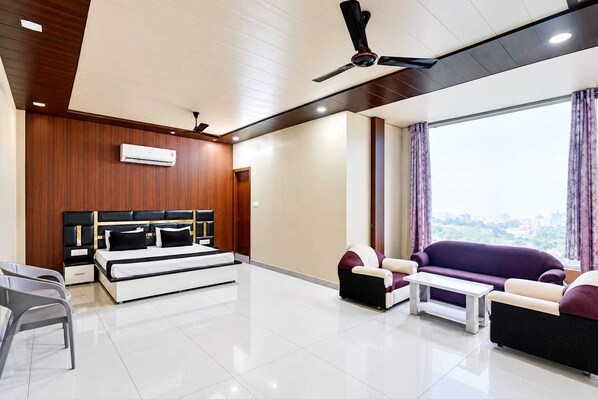 A modern living room with a sofa set, a bed with a headboard, a ceiling fan, and a large window with curtains. - Hotel O Gohana Junction Rohtak (Gohana)