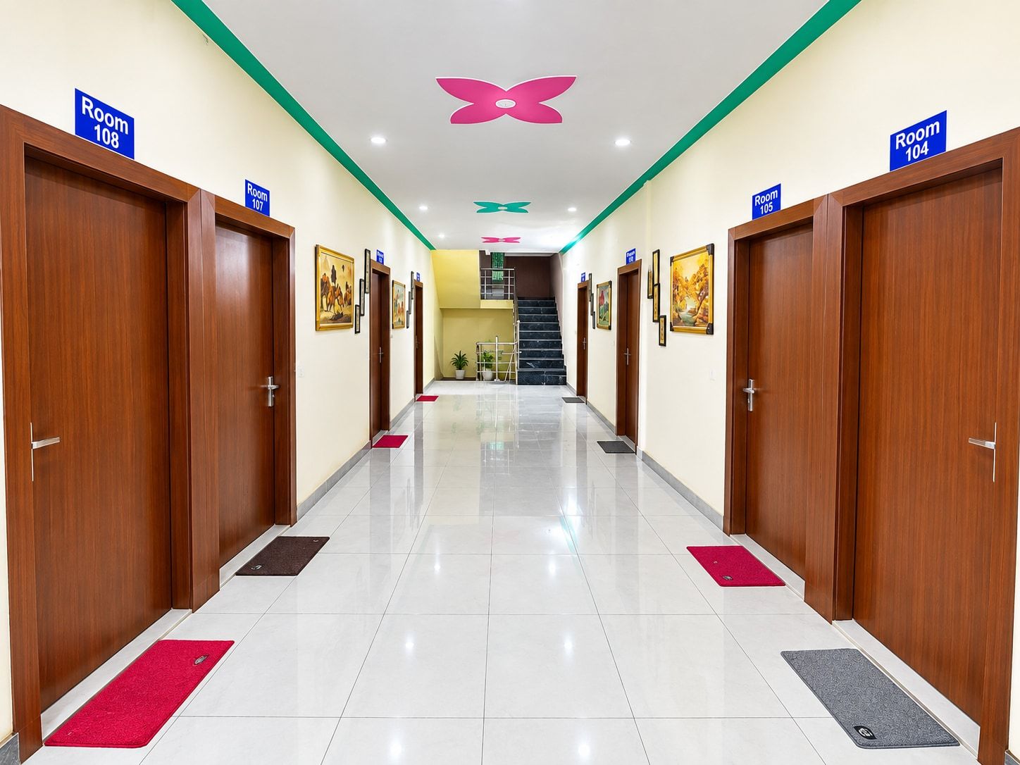 Corredor de hotel com portas identificadas como 'Room 108' e 'Room 104', uma escadaria e um ornamento de teto em formato de borboleta.