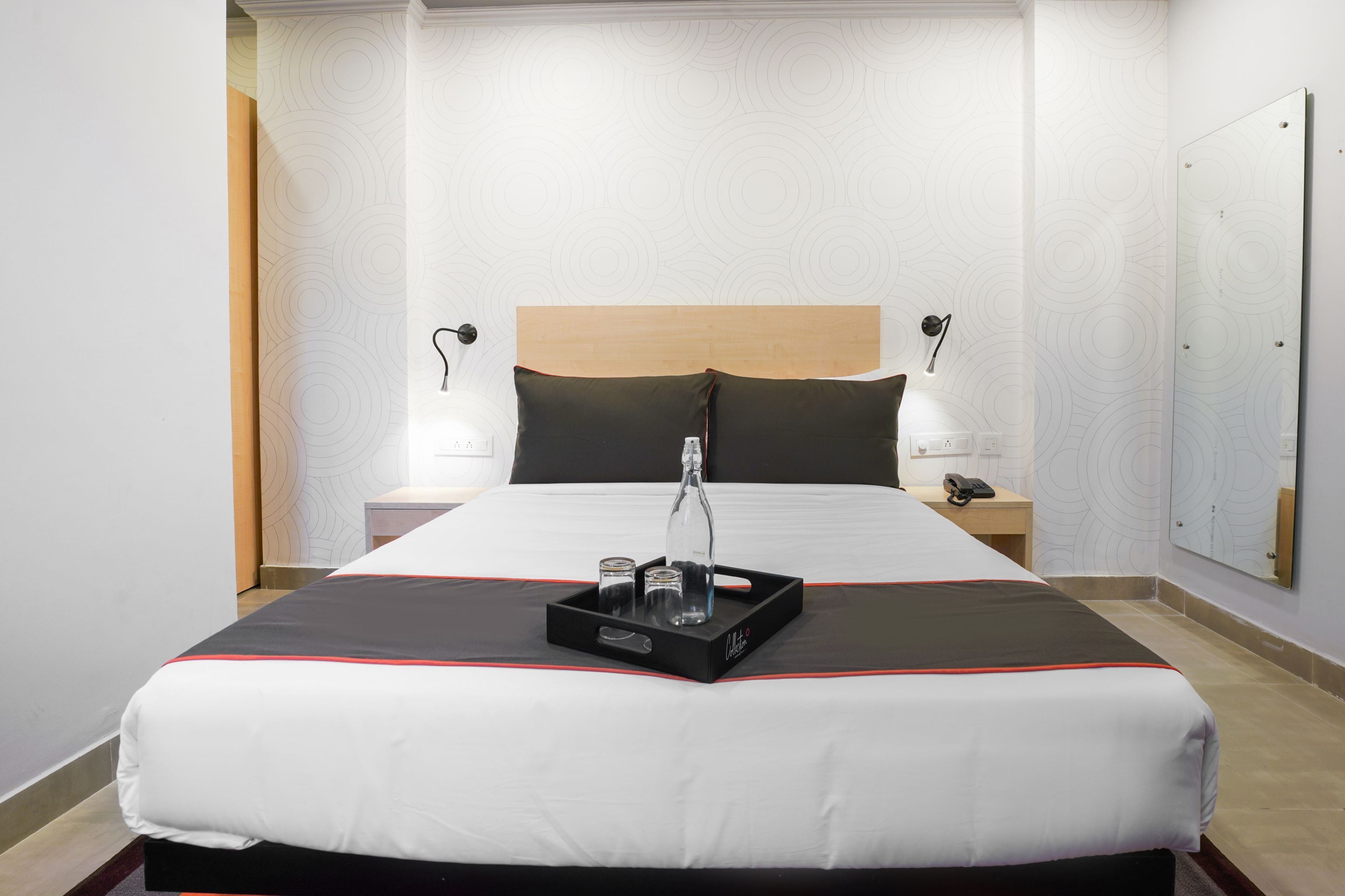 Habitación de hotel con una cama grande, mesitas de noche, un tocador, un espejo y una bandeja con una botella y vasos.