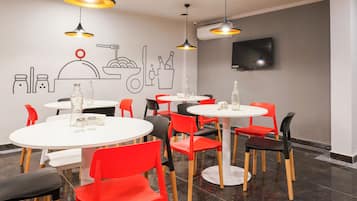 Un espace repas moderne avec des tables et des chaises rondes, un téléviseur fixé au mur et un ventilateur de plafond.