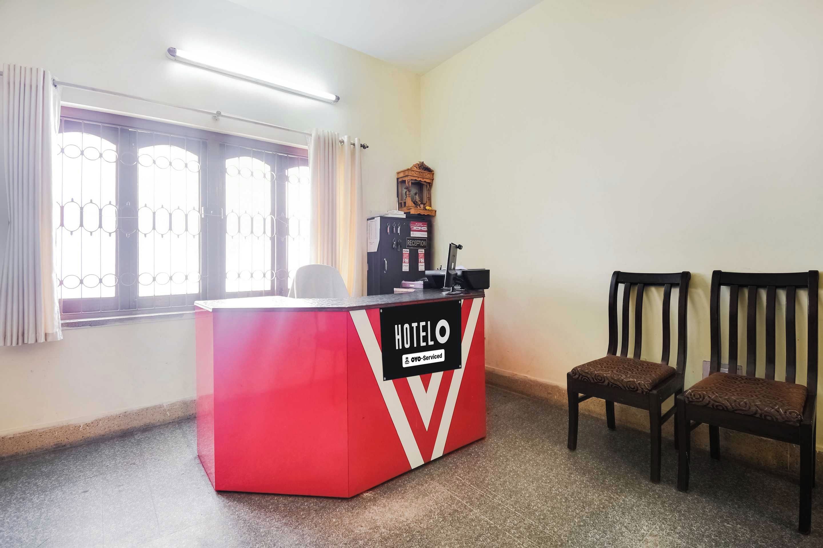 Un comptoir d’accueil aux couleurs rouge et blanc, deux chaises en bois, une fenêtre avec des rideaux et une horloge fixée au mur.