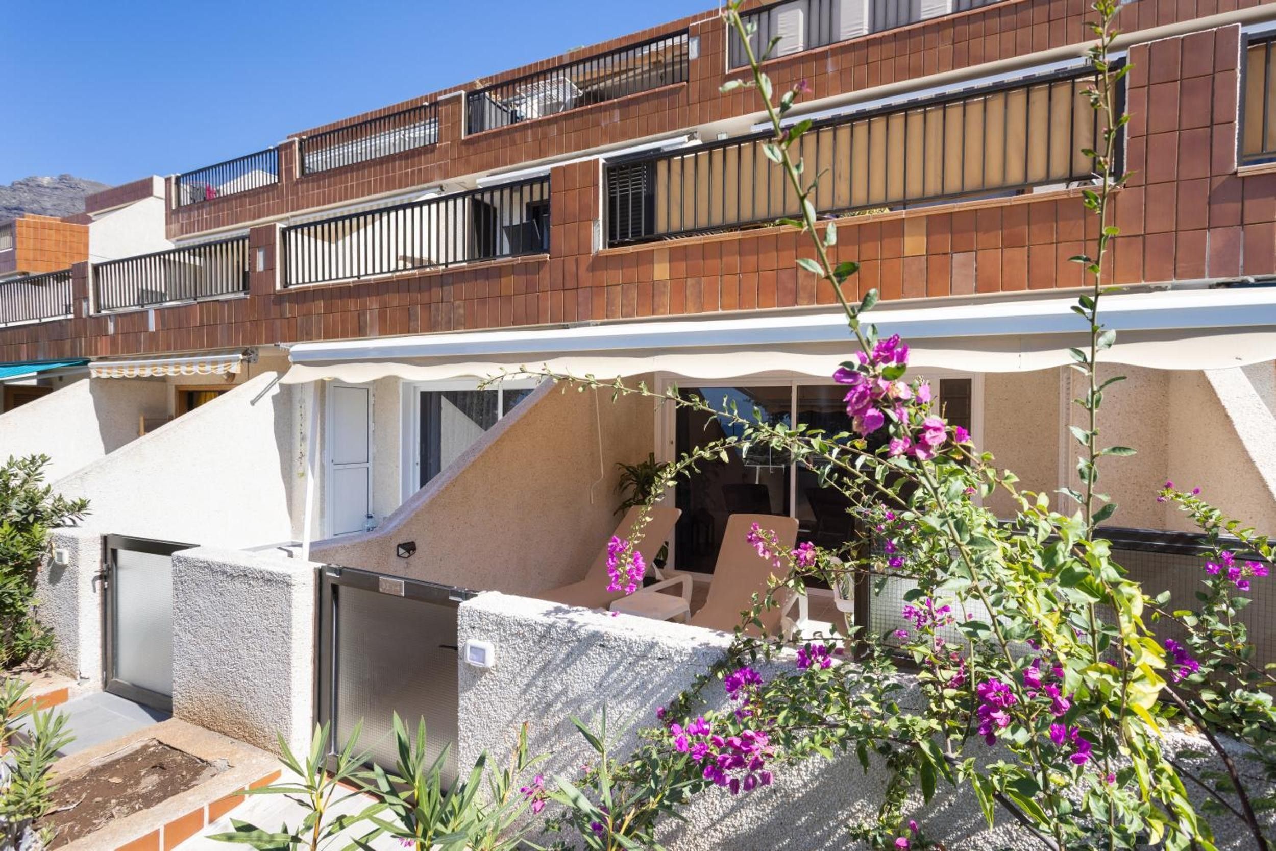 Un edificio residencial con balcón, zona de estar cubierta al aire libre y jardín con flores moradas.