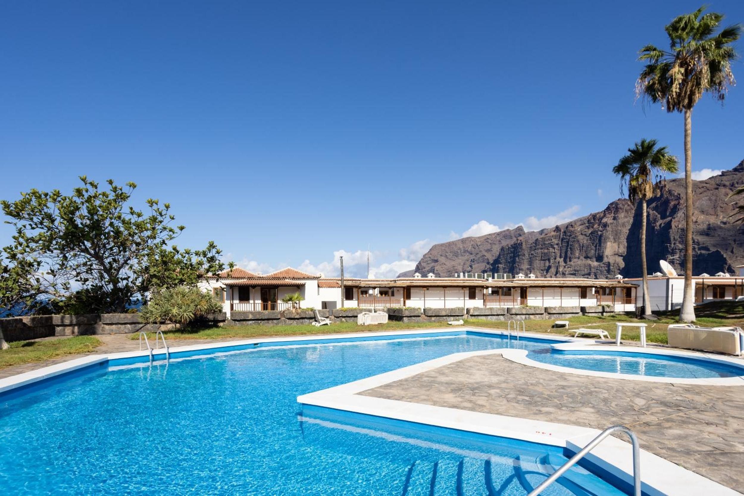 Ein Swimmingpool mit klarem, blauem Wasser, umgeben von einer Liegefläche. Im Hintergrund sind Gebäude im traditionellen Baustil und ein Bergpanorama zu sehen.