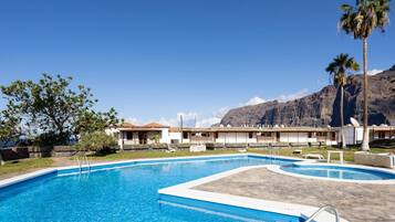 Ein Swimmingpool mit klarem, blauem Wasser, umgeben von einer Liegefläche. Im Hintergrund sind Gebäude im traditionellen Baustil und ein Bergpanorama zu sehen.