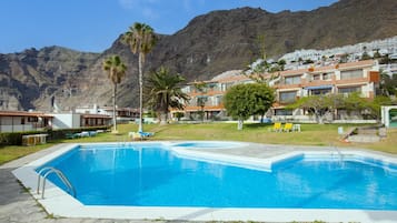 En swimmingpool med kristallklart blått vatten, omgiven av en stenbelagd terrass. I förgrunden finns solstolar och en palm. I bakgrunden syns byggnader med balkonger och ett bergigt landskap.