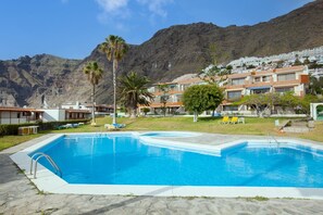 En swimmingpool med klart, blåt vand, omgivet af en belagt terrasse. Loungestole og et palmetræ ses i forgrunden. I baggrunden er der bygninger med altaner og et bjergrigt landskab.