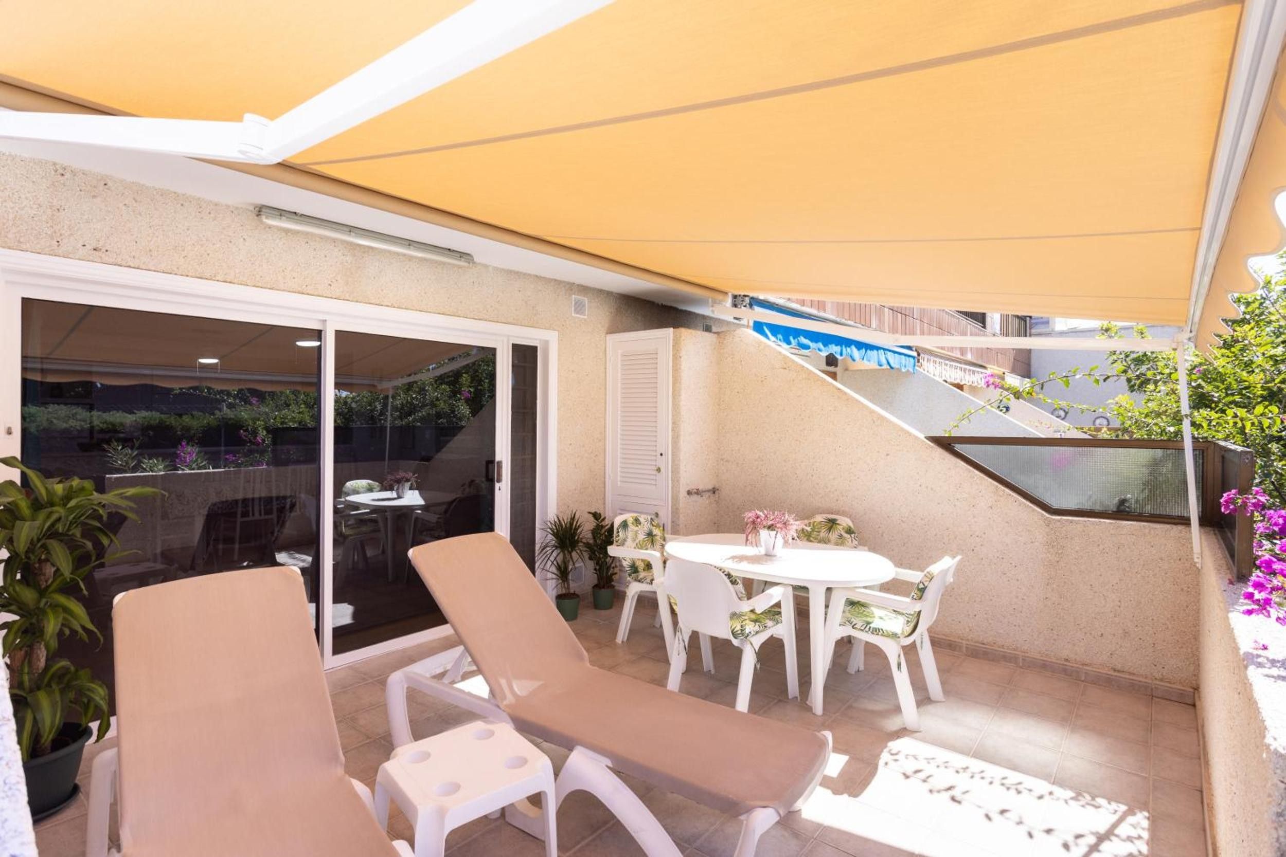Ein überdachter Außenbereich mit Lounge-Möbeln und einem Tisch, eine Glastür zum Patio und ein kleiner Garten mit Blumentöpfen.