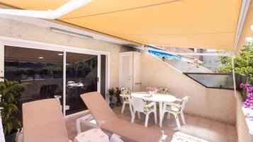 Ein überdachter Außenbereich mit Lounge-Möbeln und einem Tisch, eine Glastür zum Patio und ein kleiner Garten mit Blumentöpfen.