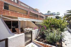 En boligbygning med en overdækket terrasse, udendørs møbler og en have med blomstrende planter.
