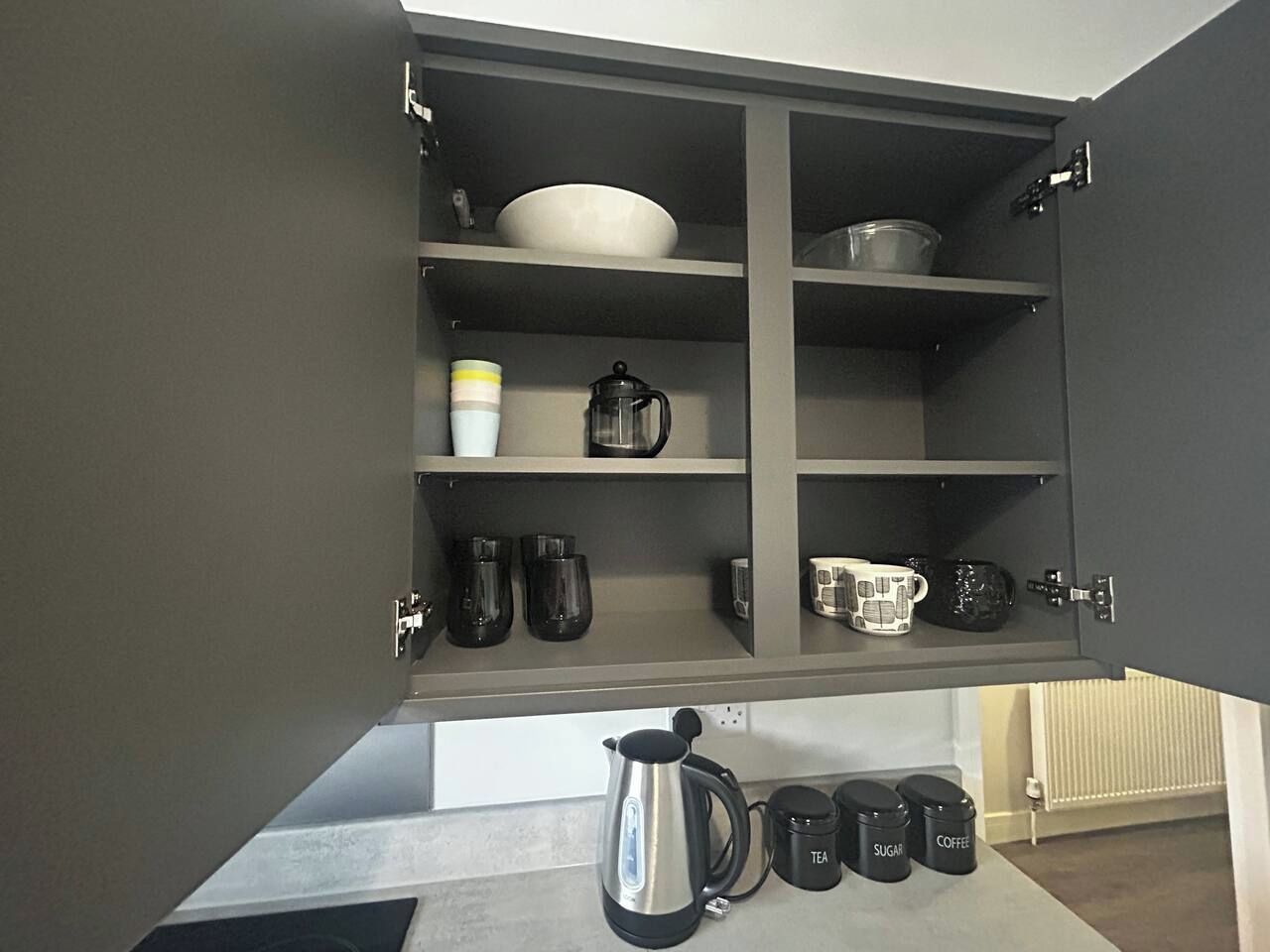 A kitchen cabinet with shelves containing various kitchenware, including a kettle, coffee pots, and mugs.