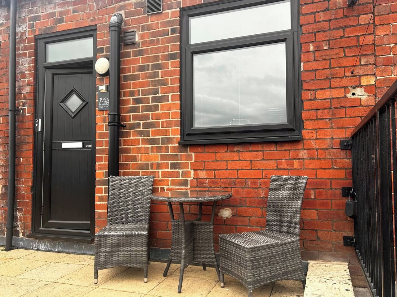 A brick building with a black door and windows, two wicker chairs, and a small round table.
