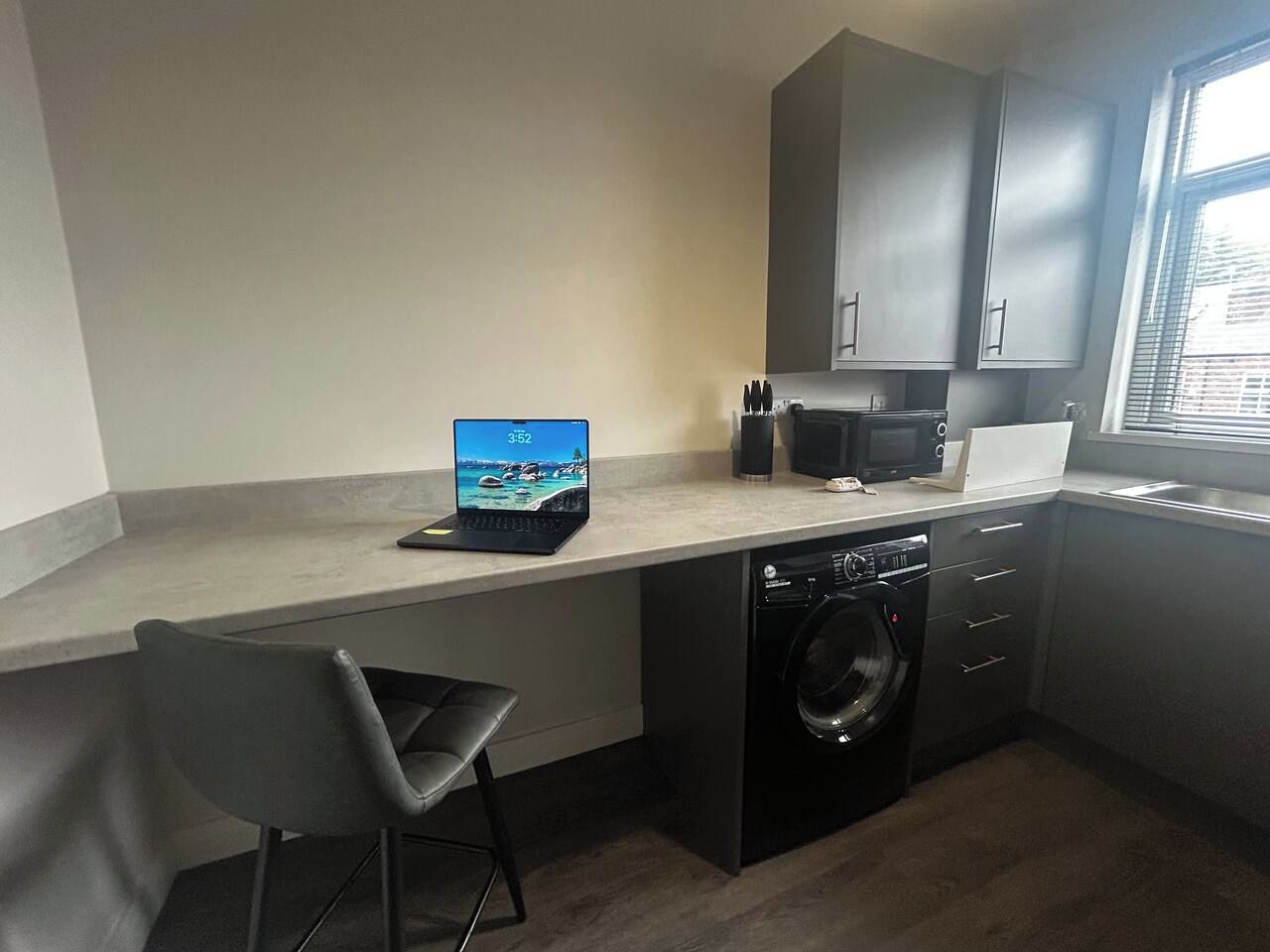 A compact kitchen with a washing machine, a laptop on the counter, and a chair.