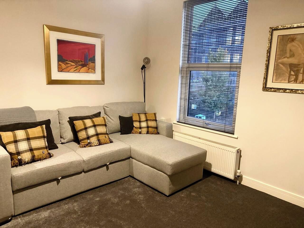 A corner living room with a grey sectional sofa, a radiator, and two framed artworks on the walls.