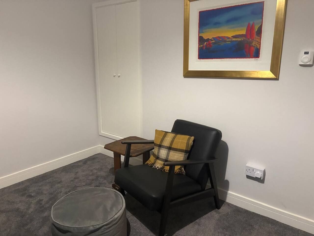 A room with a black leather chair, a small wooden table, a grey ottoman, a white door, and a framed painting on the wall.