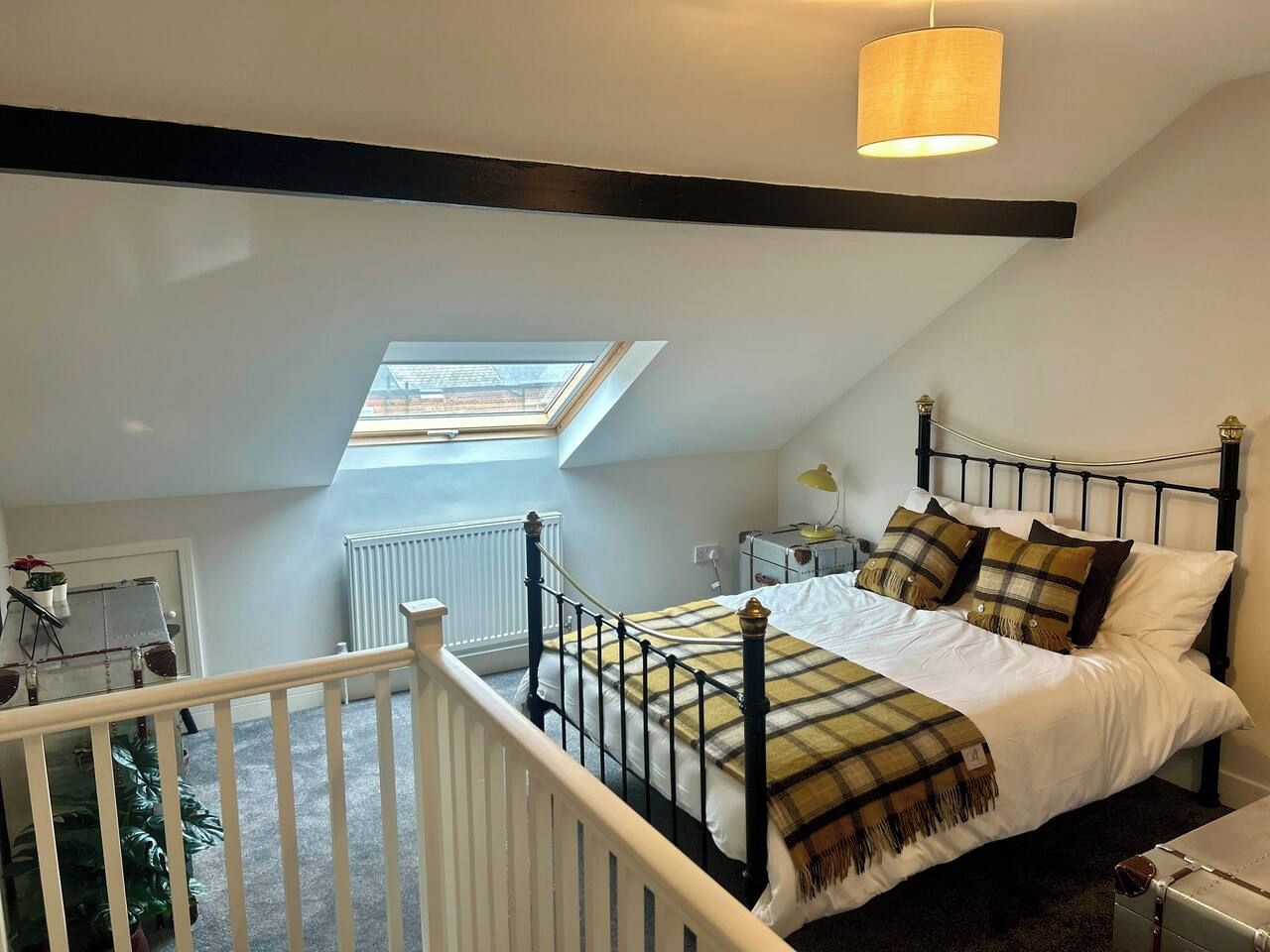 Une chambre avec un plafond en pente, un puits de lumière, un lit recouvert d’une couverture à carreaux, une table de chevet et une lampe.