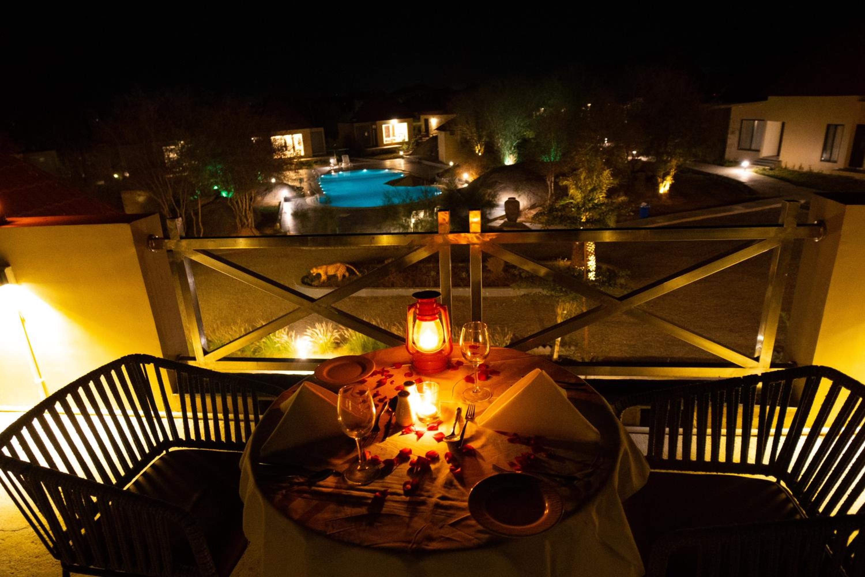 A dining table with chairs, a candle, and a glass of wine, overlooking a swimming pool and a building.