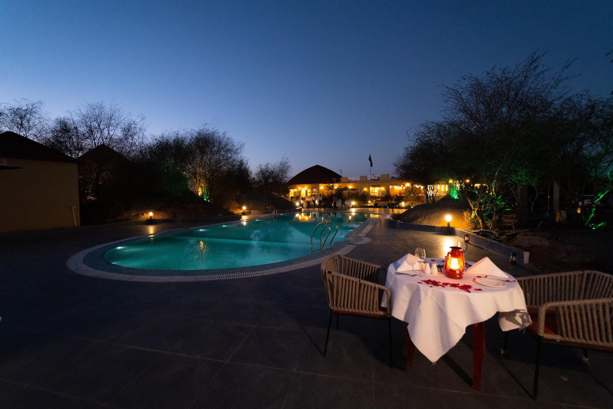 An outdoor swimming pool area with a table set for dining, surrounded by chairs, and a building in the background.