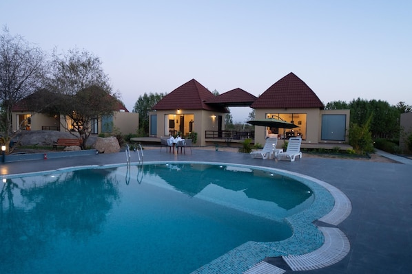 A swimming pool with lounge chairs and a covered seating area, surrounded by buildings with red roofs. - Jawai Zinc Journey By The Fern, Jawai (Pali)