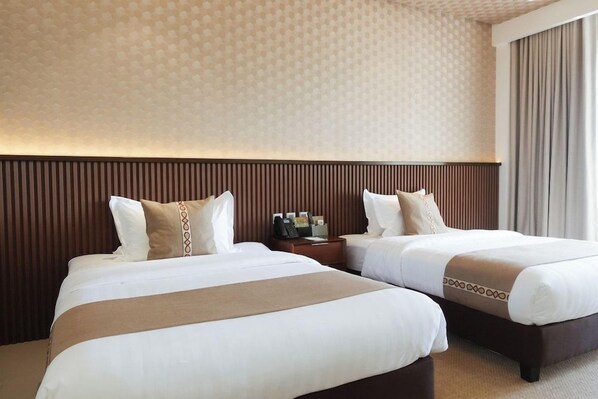 Habitación de hotel con dos camas, cabecera de madera, mesita de noche con lámpara y ventana con cortinas.